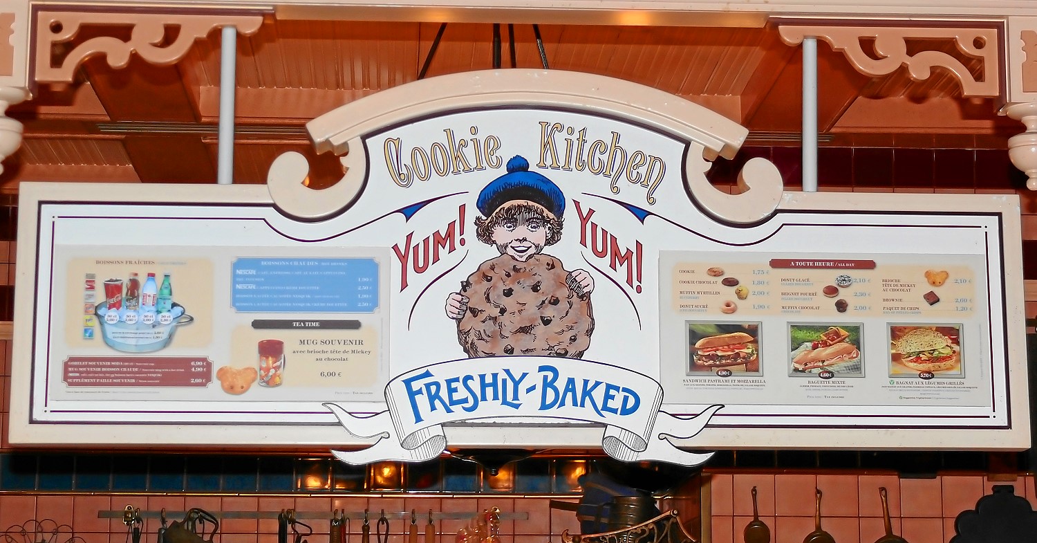 Cookie Kitchen Cookie Kitchen sign above the counter with a child biting into a giant cookie. To the left and right of it, the counter's offer is pointed out