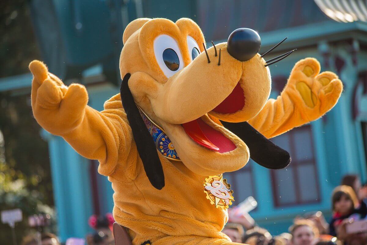 Pluto with outstretched arms Pluto is sitting on the back of a parade float with his arms spread wide. Instead of his usual dog tag, he has a pendant with a large 25 tied around his neck