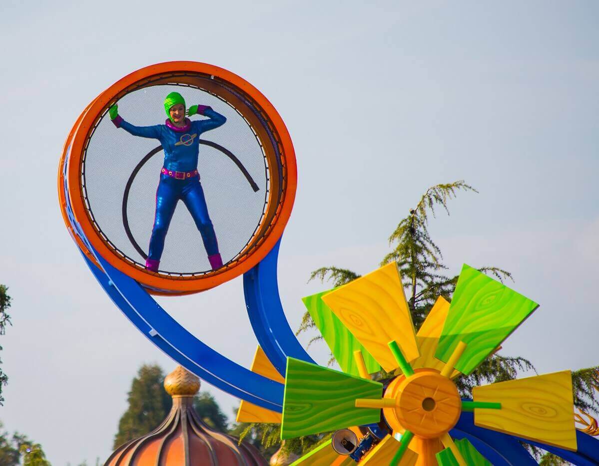 acrobat on the parade float An actobate in a blue suit and green cap stands in a kind of cage at a lofty height, far above the parade float