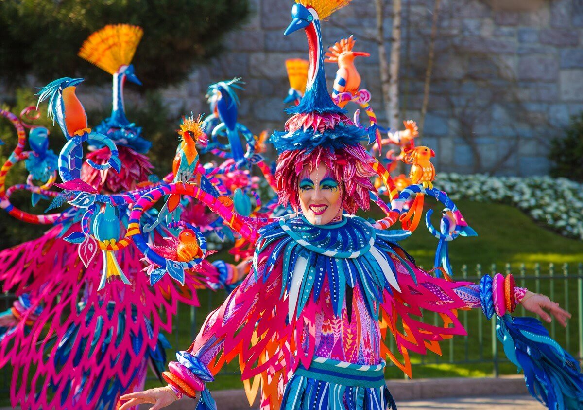 Dancer as bird creature A group of dancers are dressed as bird creatures. The costumes are covered with feathers and on the head and shoulders are various small birds