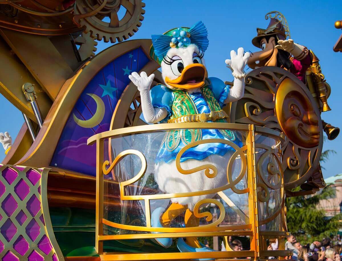 Daisy Duck at the parade Daisy Duck is standing in a pulpit on the parade float. She is wearing a steampunk outfit with light blue and green elements. Goofy can also be seen on the back of the float