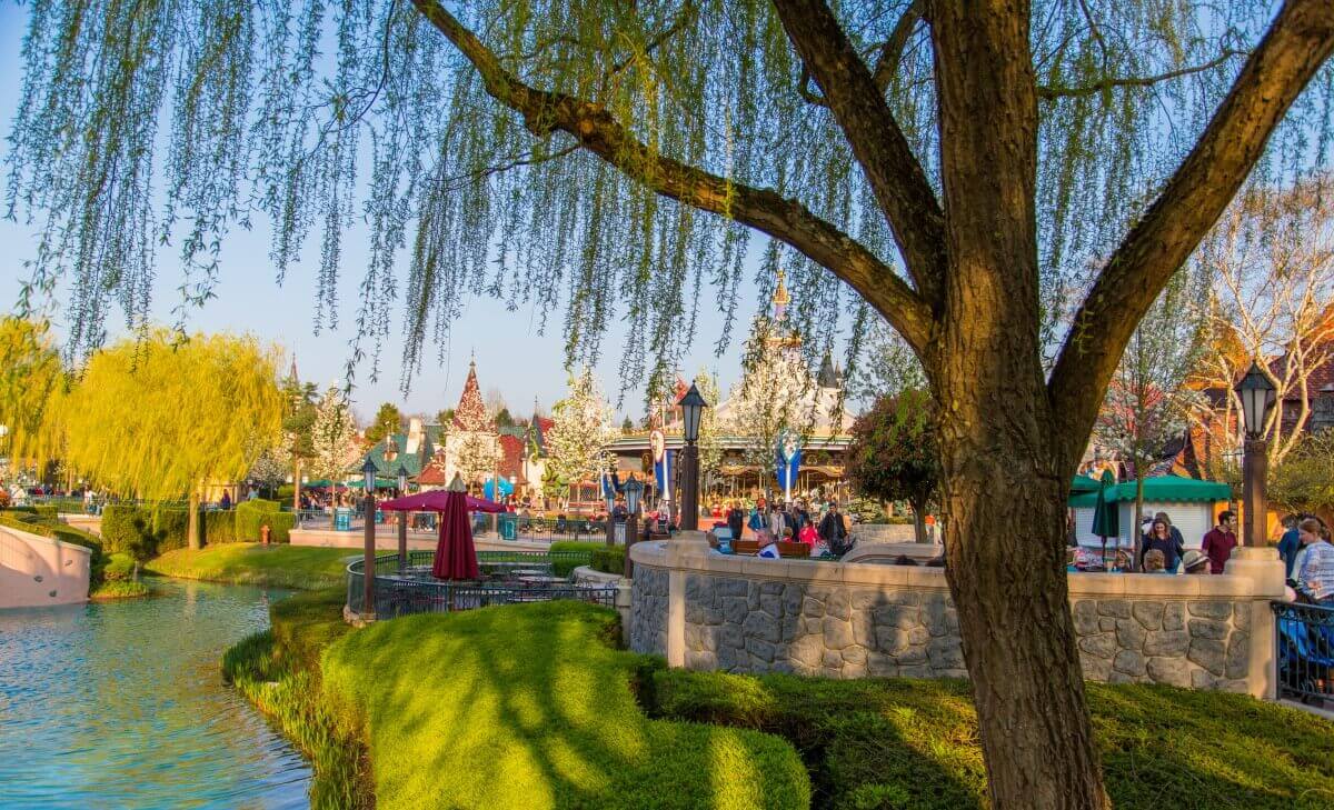 Idyllic design of the Fantasyland behind a big tree in the foreground green areas, a path and seats of a fast food restaurant are visible