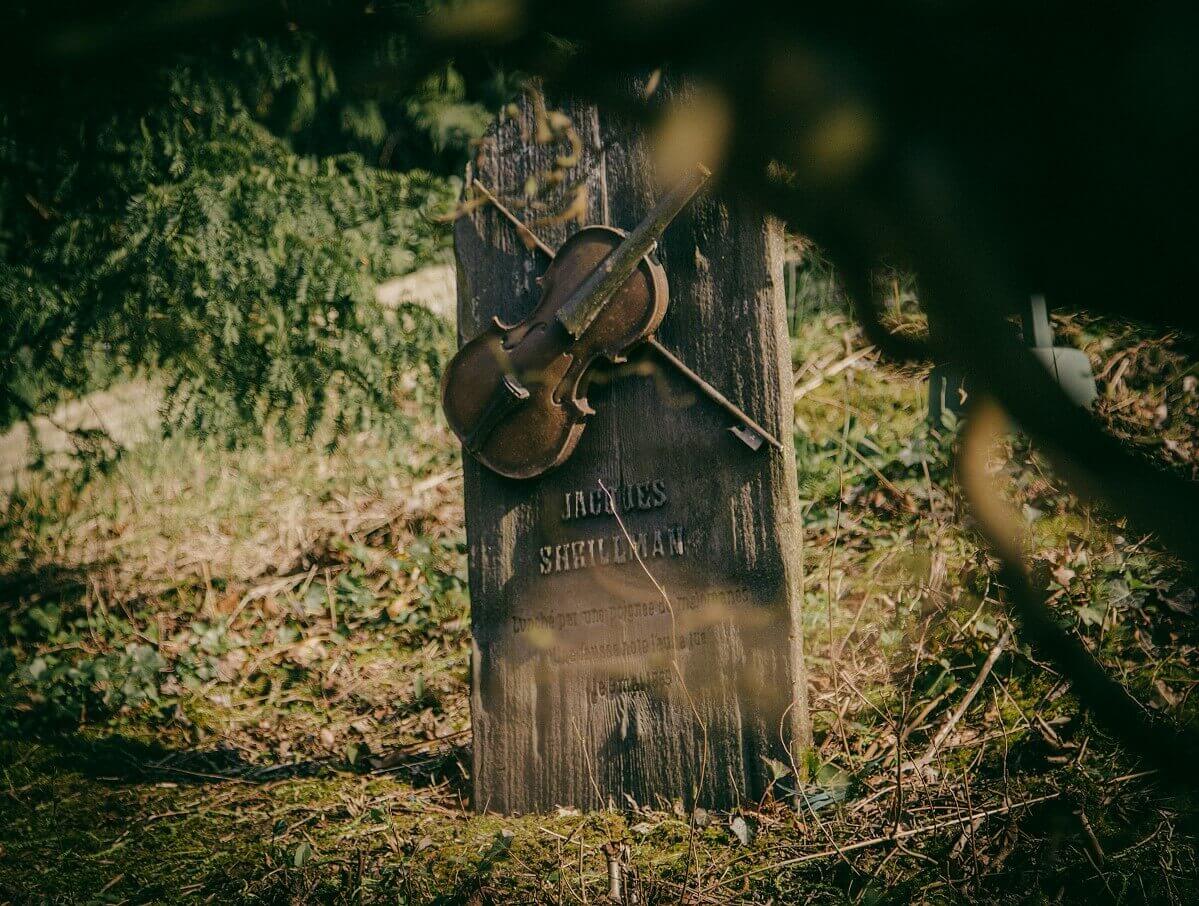 Gravestone at Boot Hill A view, slightly obscured by branches, of a gravestone with a violin and the inscription Jacques Shrillman on Boot Hill.