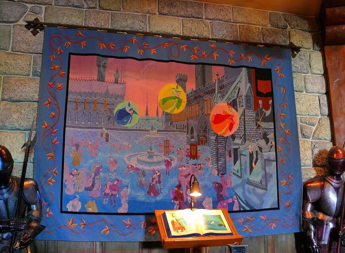 Tapestry with the three fairies A large tapestry shows the castle courtyard over which the three fairies hover, the carpet is framed by two knights armor