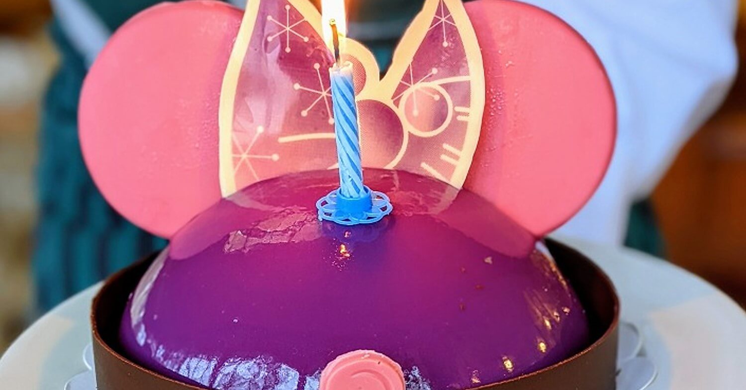 Disney birthday cake A purple frosted cake with a blue candle in the center and two pink chocolate stars arranged like Mickey Mouse ears is served on a white plate