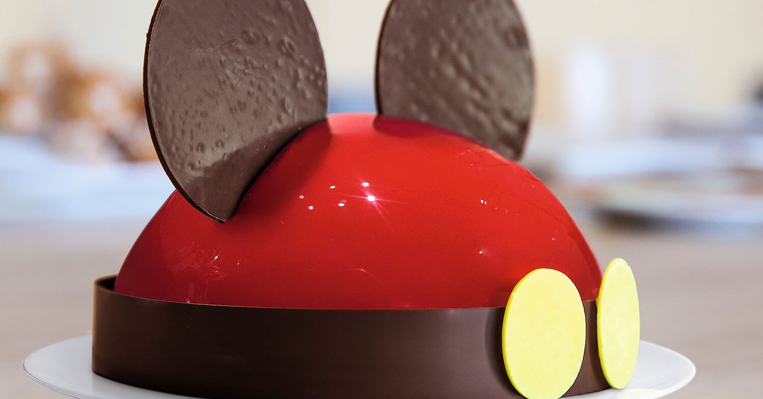 Mickey Mouse cake A cake with the look of Mickey Mouse. A red iced cake with a band of chocolate with two yellow thalers attached to the front. On top of the cake are two large chocolate wafers to represent the ears of Mickey.