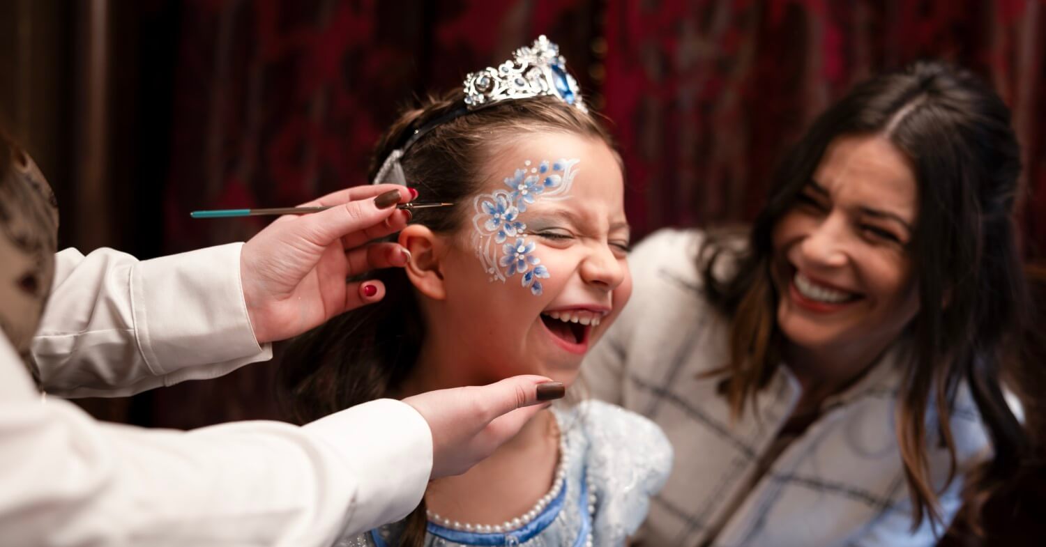 My Royal Dream A little girl is wearing a silver tiara and make-up on her face. She is laughing, just like her mother, who is sitting next to her