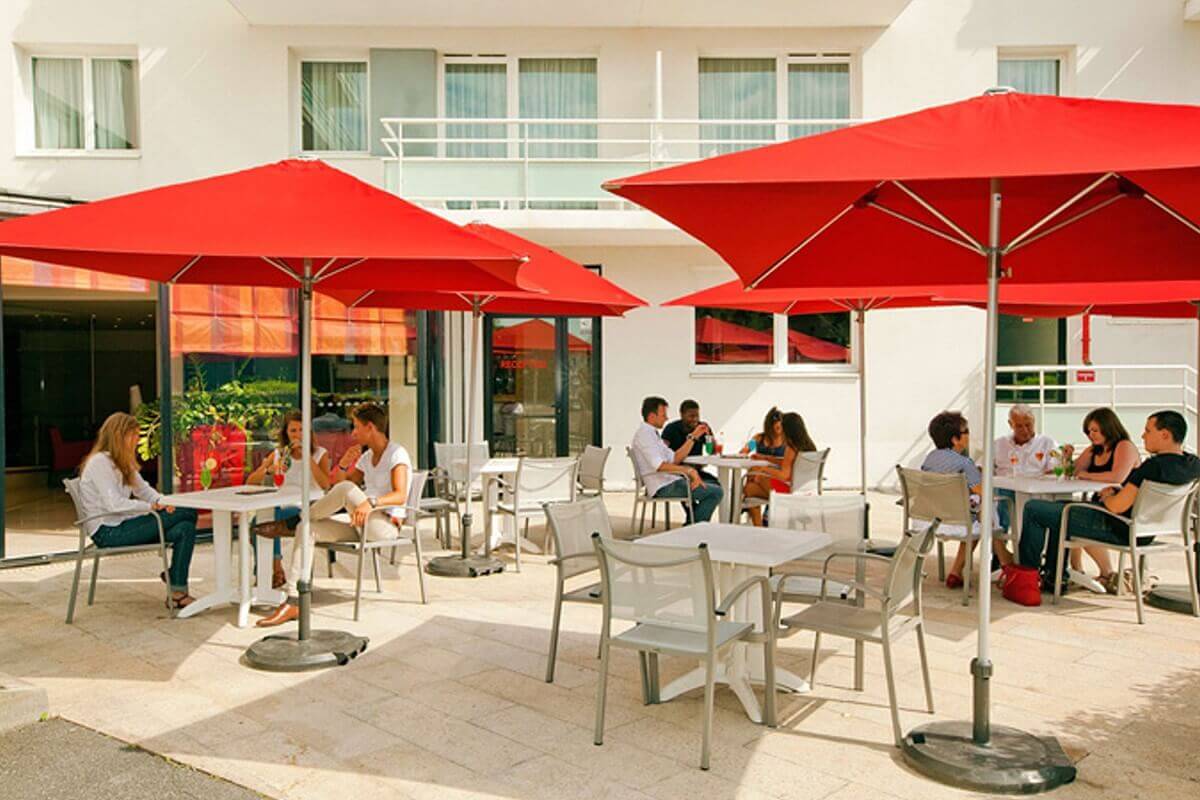 Terrace Terrace in front of the Residhome Hotel, white tables with four grey chairs each are covered by red sunshades, some tables are occupied by guests