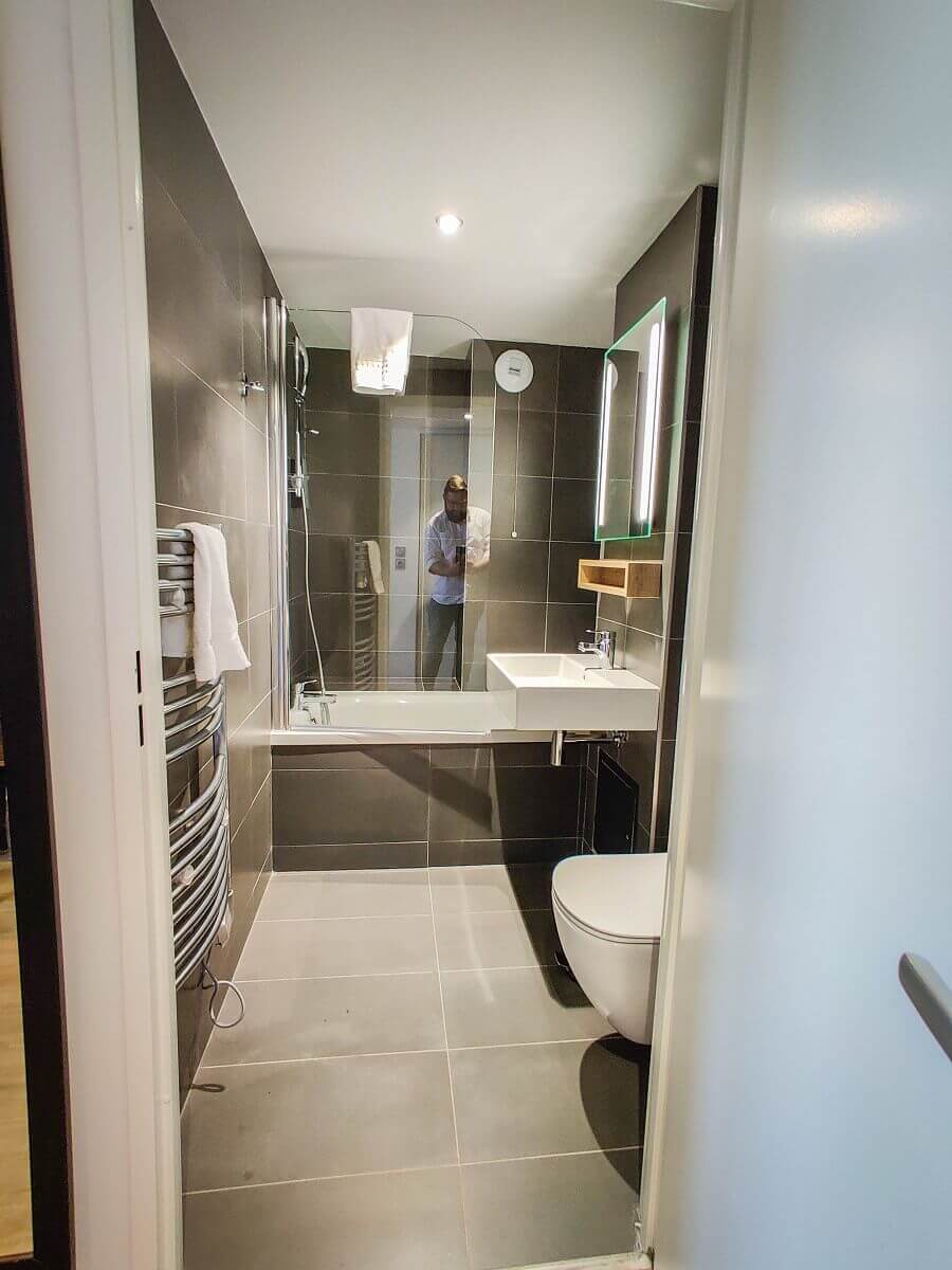 Bathroom View into the hotel bathroom with dark grey tiles, white washbasin and white WC as well as a shower tray